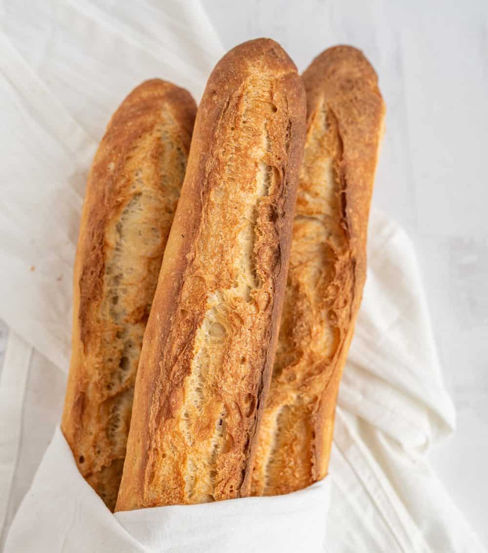 Three crusty French baguettes, made from a classic baguette recipe, are wrapped partially in a white cloth and rest on a white surface.