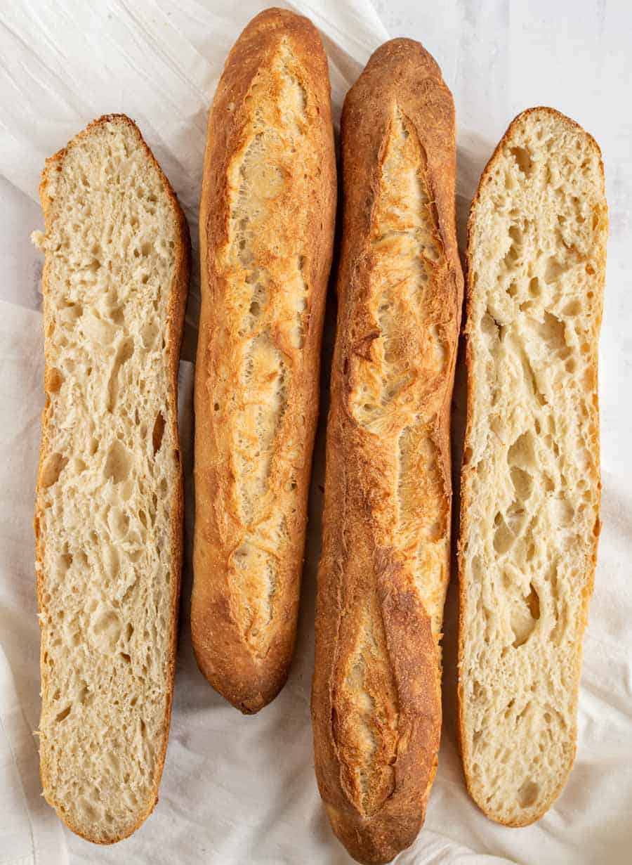 Four baguettes are arranged side by side on a white cloth; two are shown whole, and two are cut lengthwise to reveal their airy, textured interior crumb—a perfect showcase for any classic baguette recipe.
