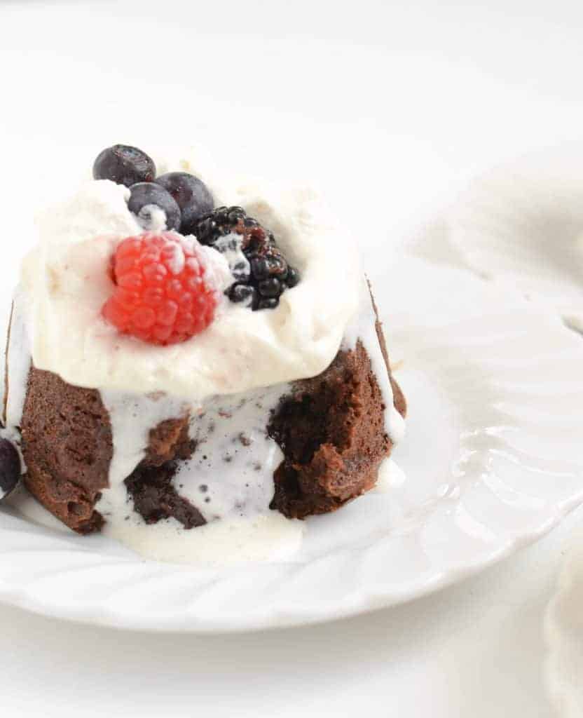 Side view of a drippy lava cake that has been opened and the center is running out whipped cream and berries on top.