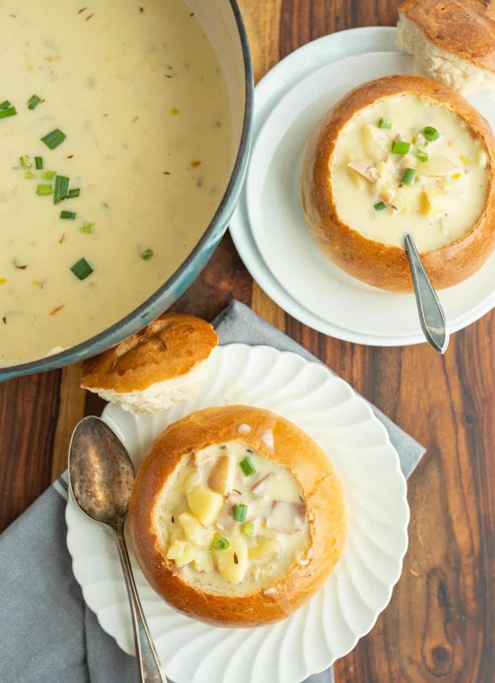 top view of a creamy clam chowder with canned clams soup and two plates next to it with crunchy round bread bowls on white plates with the top perfectly cut off and chunky cream soup and a spoon.