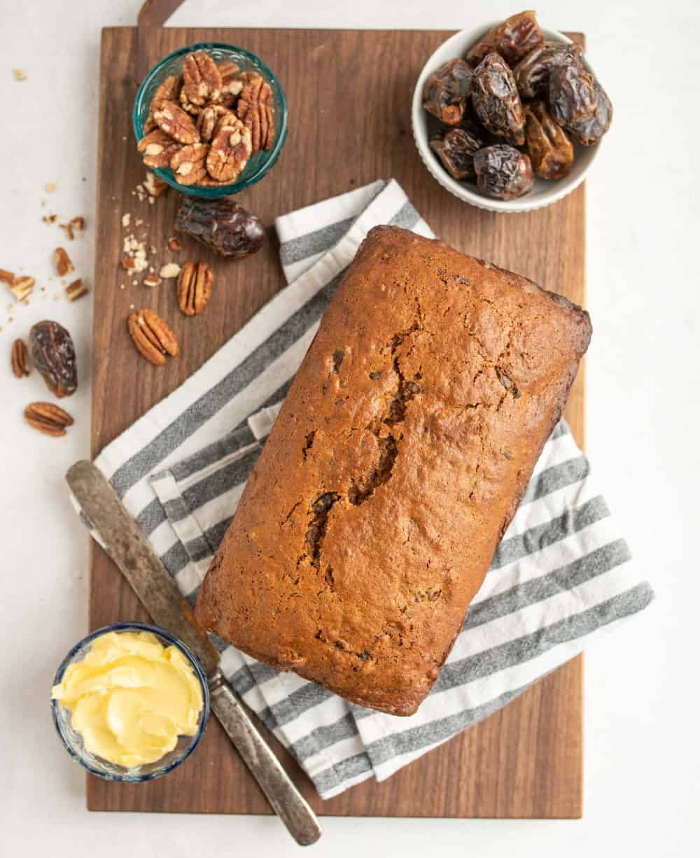 Top view of an unsliced loaf of date nut bread on a striped napkin and cutting board with ramekins of butter, pecans, and dates next to it.