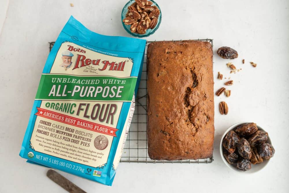 Top view with a white background on a cooling rack is a loaf of cinnamon colored perfectly baked loaf with a crack in the middle with nuts and dates and flour all around.