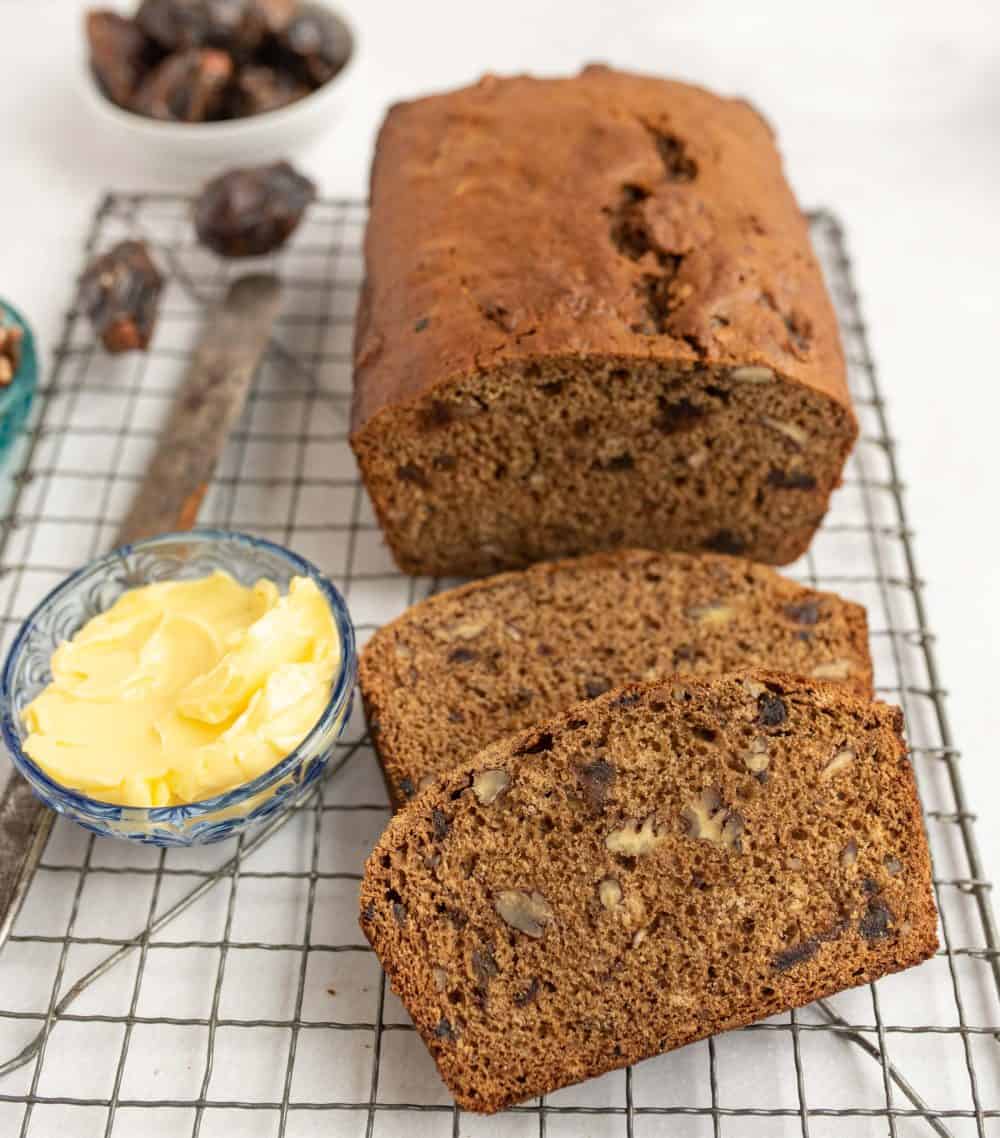 Close up of slices of date nut bread on a cooling rack next to a ramekin of butter and the rest of the unsliced loaf.