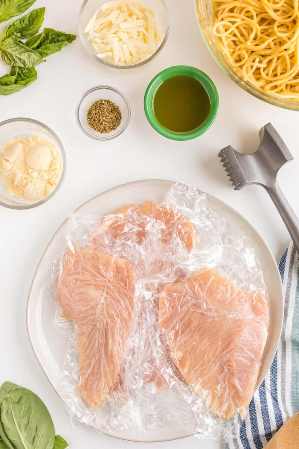Top view of all the starting ingredients for parmesan chicken including the raw chicken in cling wrap and a meat pounder, spices and cheese in dishes around the outside.