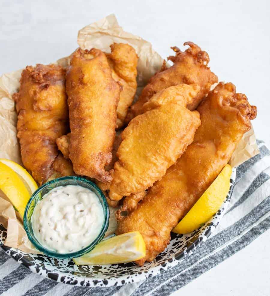 A basket of crispy golden fried fish fillets, made with a classic fish batter recipe, served with lemon wedges and a small bowl of tartar sauce, placed on a striped cloth.