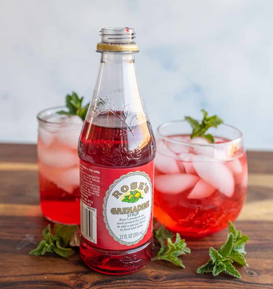 aesthetic photo of a bottle of 'Rose's Grenadine" with tumblers of ice and garnish and a red "Shirley-Temple" drink.