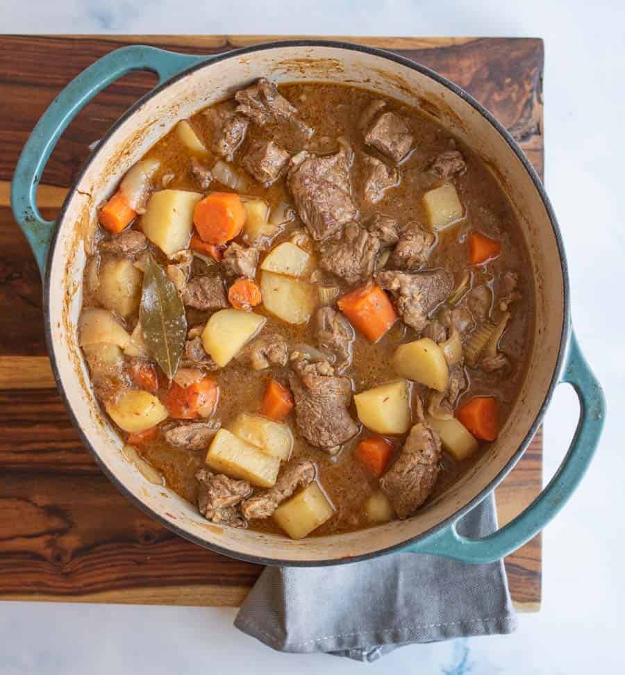 A pot of old fashioned beef stew recipe with chunks of beef, potatoes, carrots, and a bay leaf in brown broth sits on a wooden board with a gray cloth underneath.