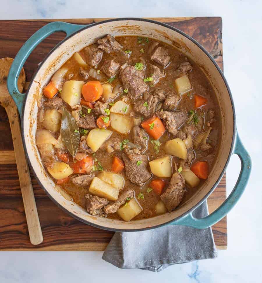 A blue Dutch oven filled with an old fashioned beef stew recipe—featuring tender beef, carrots, potatoes, and herbs—sits on a wooden cutting board beside a wooden spoon and a gray napkin.