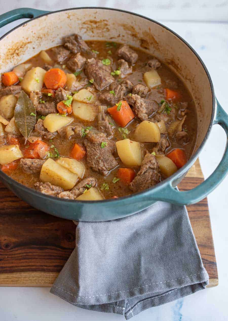 A large blue pot filled with beef stew, including chunks of beef, potatoes, carrots, and a bay leaf, sits on a wooden board with a gray cloth underneath.