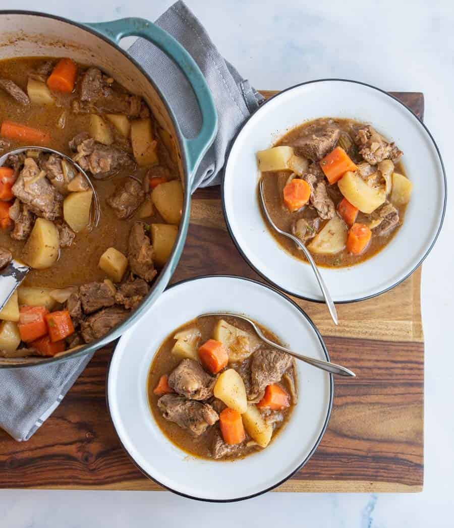 A pot of beef stew with chunks of meat, carrots, and potatoes sits on a wooden board. Two bowls filled with the stew and spoons are beside the pot, ready to eat.