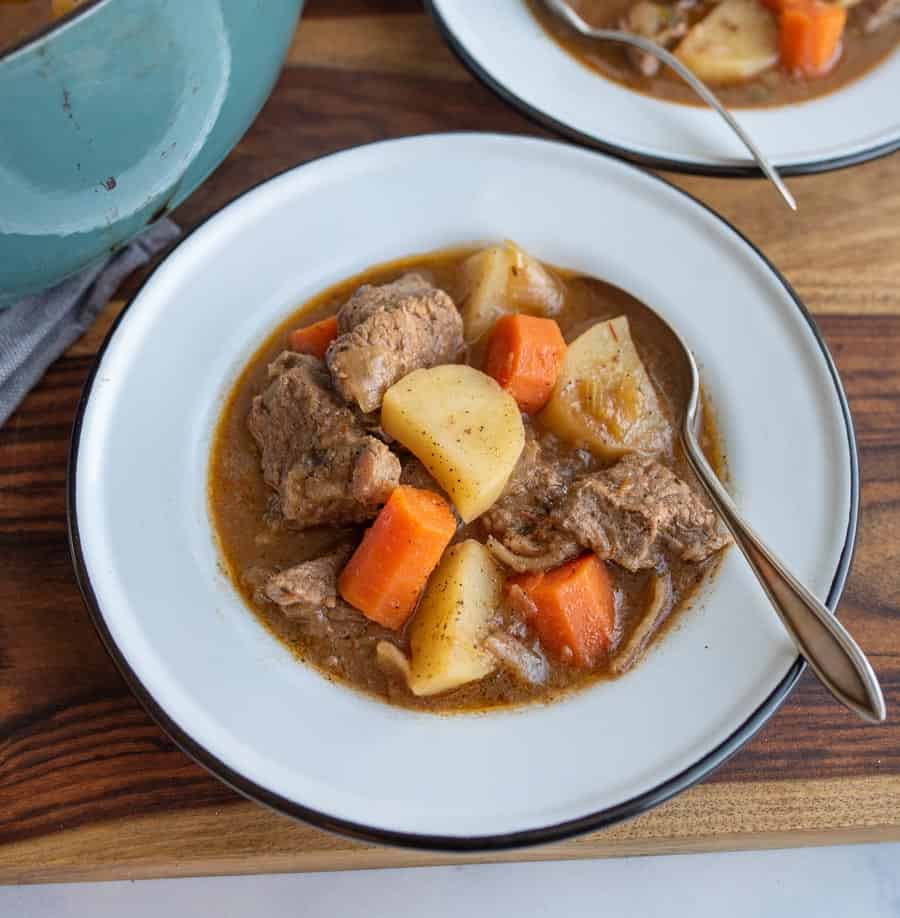 A bowl of hearty old fashioned beef stew recipe with chunks of beef, potatoes, and carrots in brown broth, served with a spoon on a white plate with a black rim, set on a wooden table.