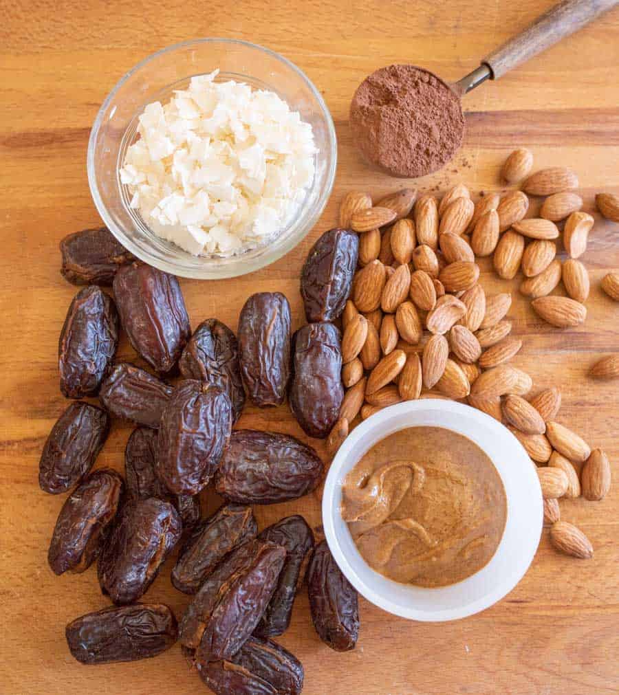 A wooden surface with whole dates, raw almonds, a glass bowl of coconut flakes, a small bowl of almond butter, and a spoonful of cocoa powder arranged neatly.