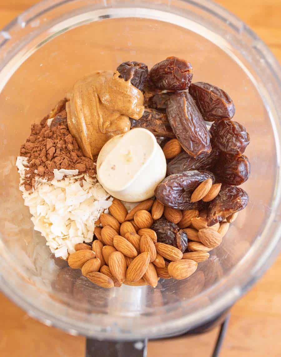 A food processor bowl filled with whole almonds, dates, shredded coconut, cocoa powder, and a dollop of peanut butter—ingredients ready to be blended into delicious and nutritious Healthy Snack Bars.