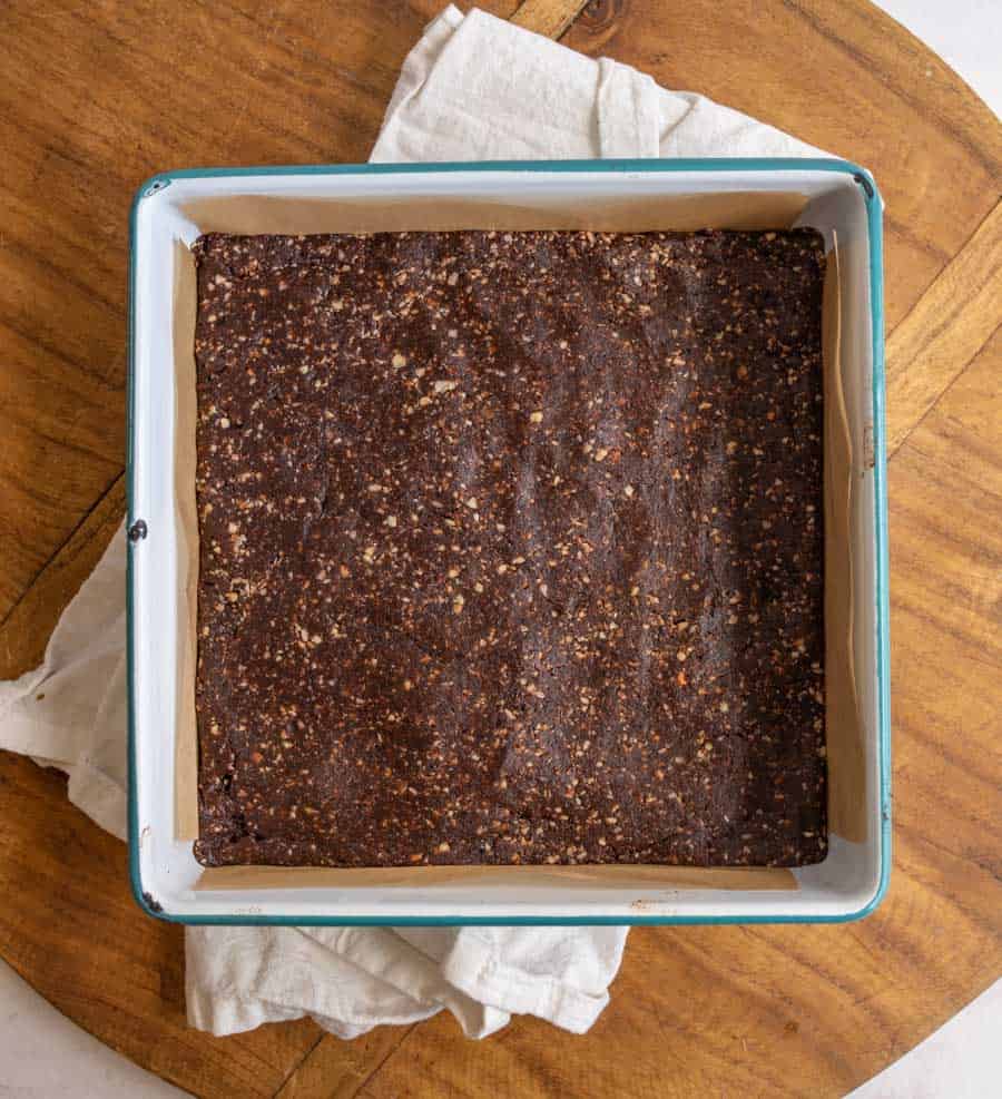 A square baking pan lined with parchment paper holds an unbaked, pressed chocolate mixture on a wooden surface with a beige cloth underneath.