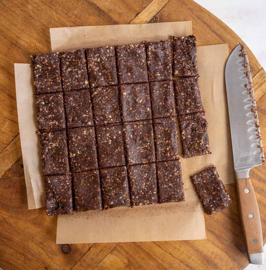 A batch of homemade healthy snack bars, cut into 24 chocolatey squares, sits on parchment paper atop a wooden table. A large serrated knife lies to the right of the bars.