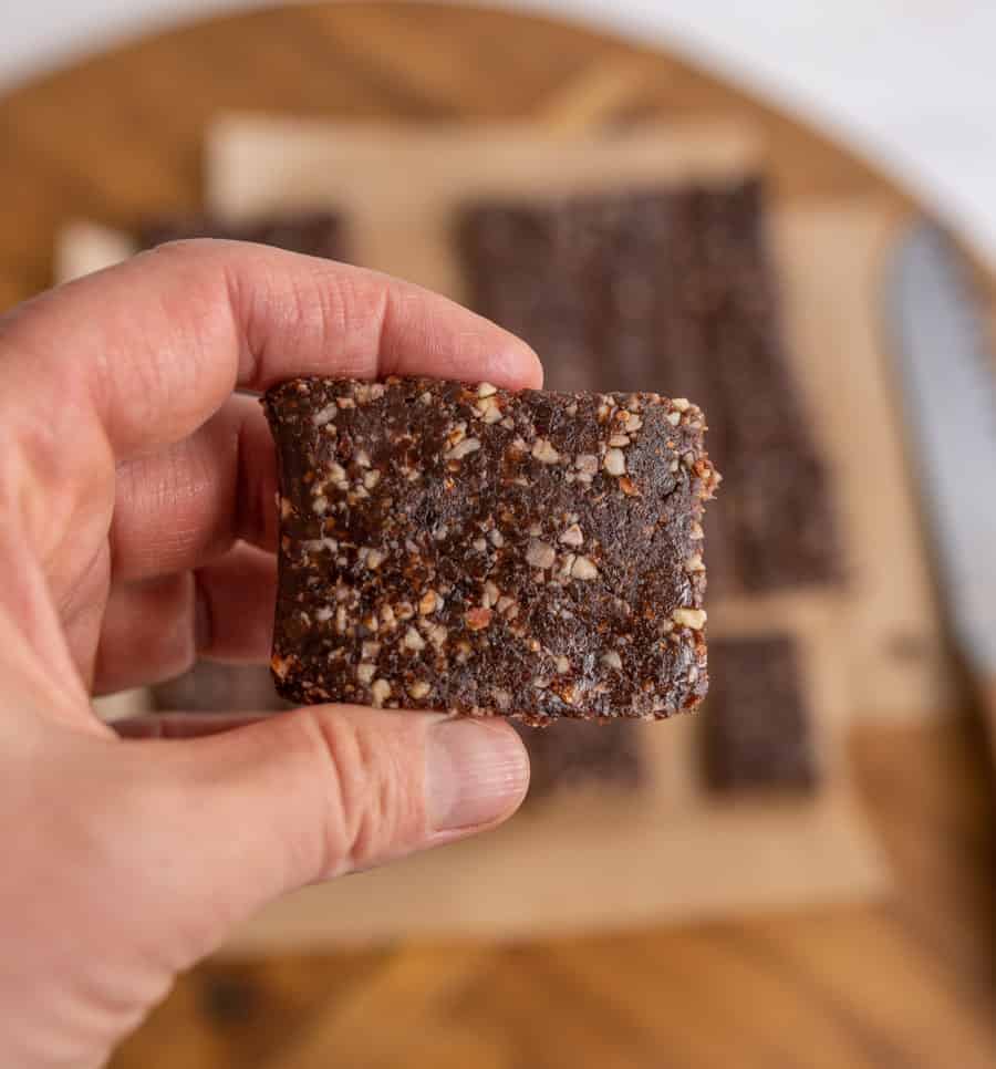 A hand holds a rectangular homemade Healthy Snack Bar with visible pieces of nuts. More bars are on parchment paper in the blurred background.