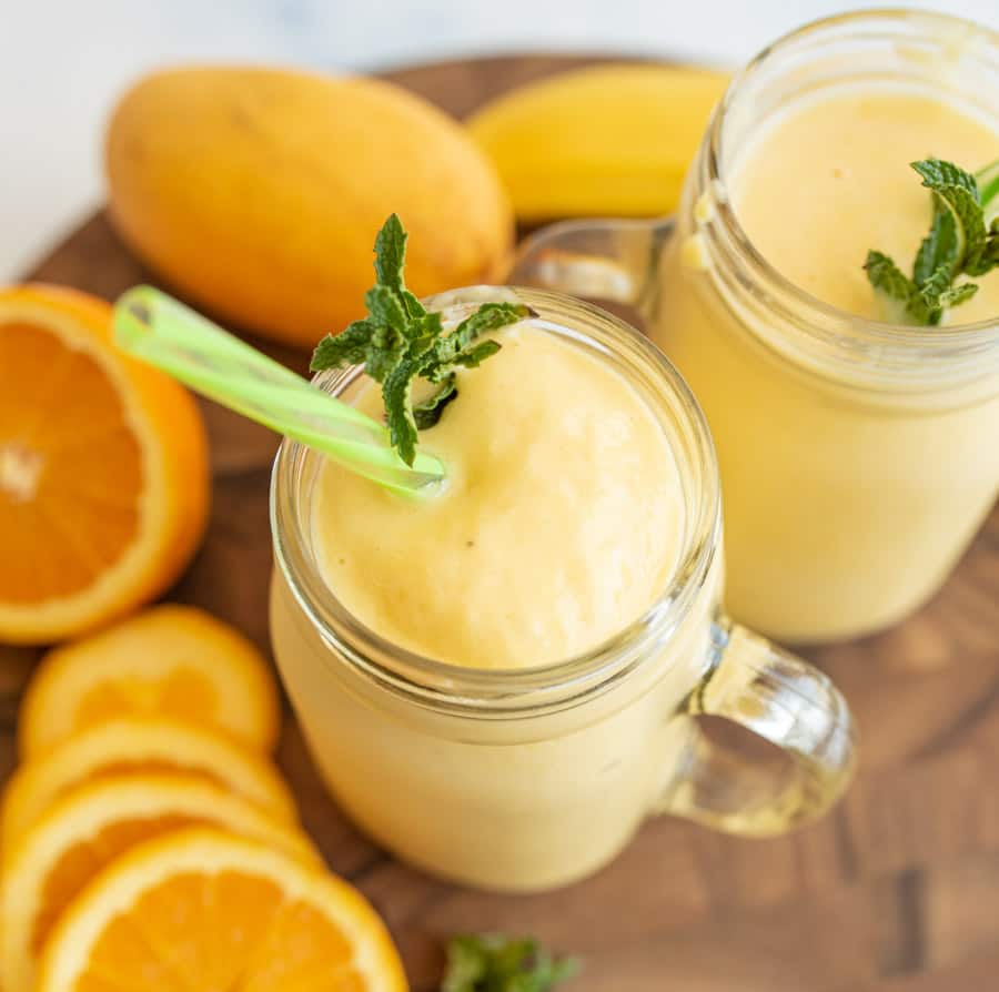 Two mason jars filled with creamy yellow smoothie, each topped with a mint sprig and a green straw. Slices of orange and a whole mango are arranged on a wooden surface beside the jars.