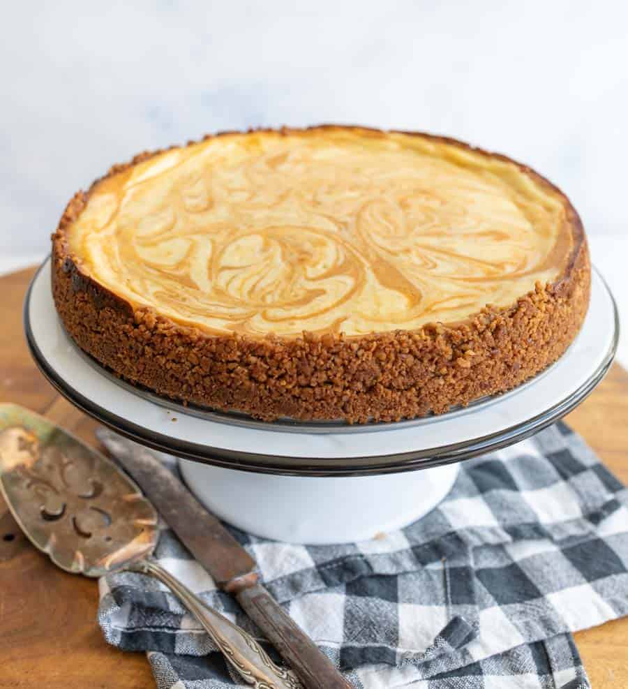 A marble-swirled cheesecake with a crumbly brown crust sits on a white cake stand. A pie server and knife rest nearby on a black and white checkered cloth on a wooden surface made from a pumpkin cheesecake recipe.