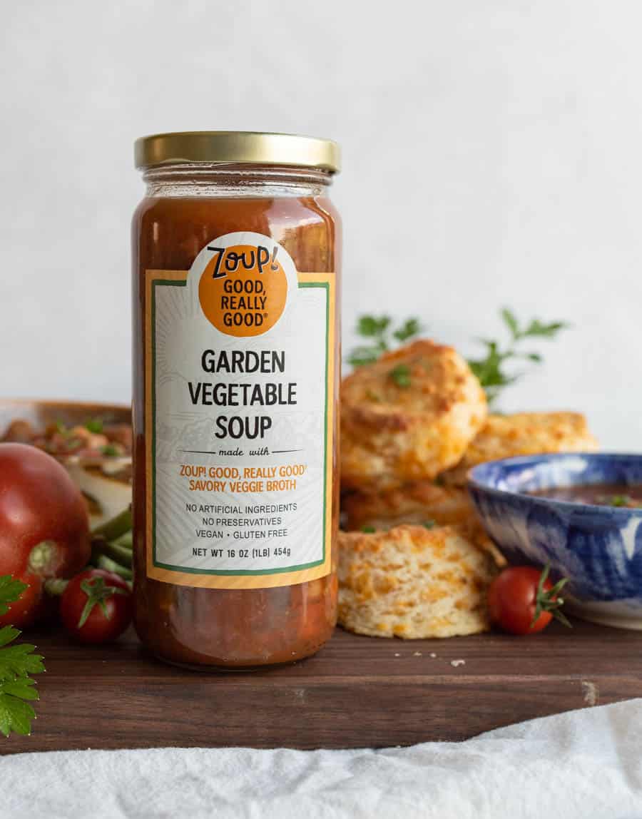 A jar of Zoup! Good Really Good! Garden Vegetable Soup stands on a white surface, surrounded by fresh tomatoes, herbs, biscuits, and a blue patterned bowl in the background.