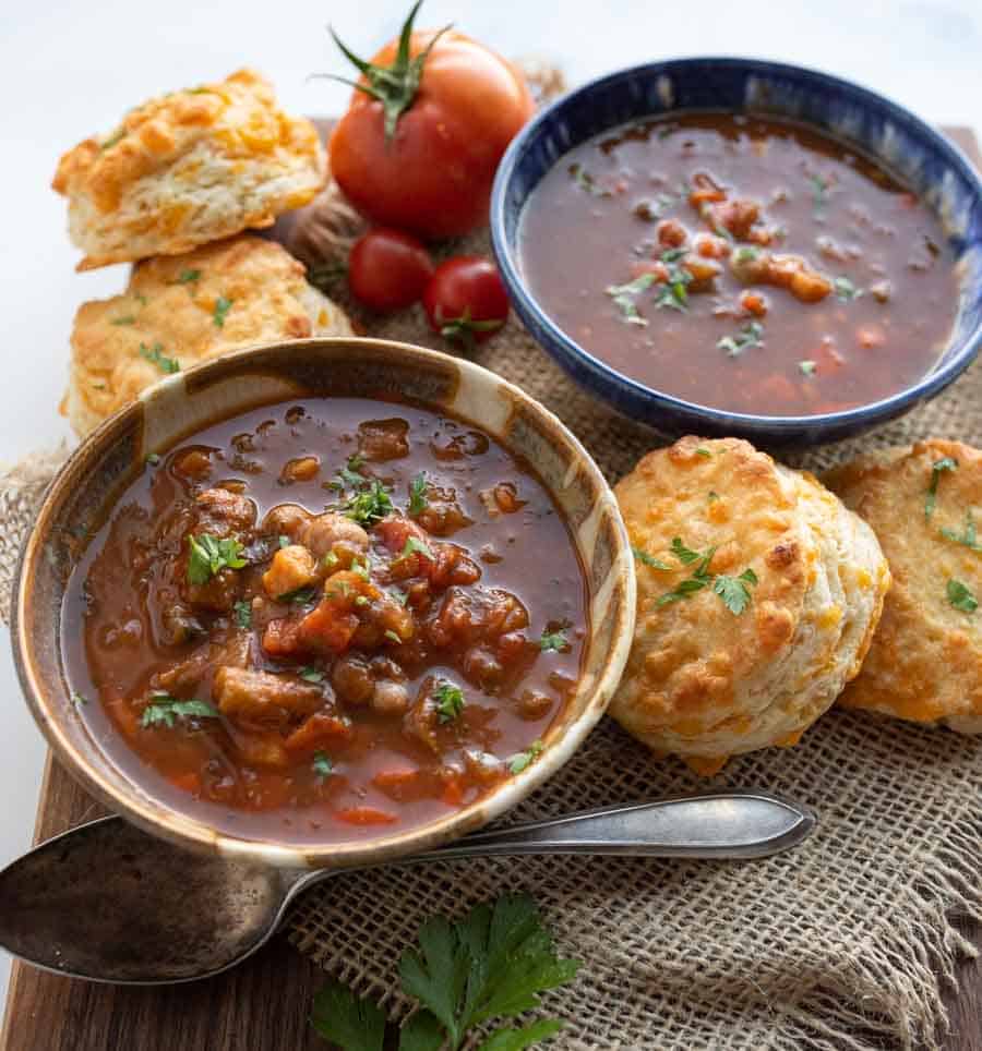 Two bowls of hearty stew garnished with herbs, surrounded by golden cheddar biscuits, fresh tomatoes, and parsley, all arranged on a rustic wooden board with burlap underneath.