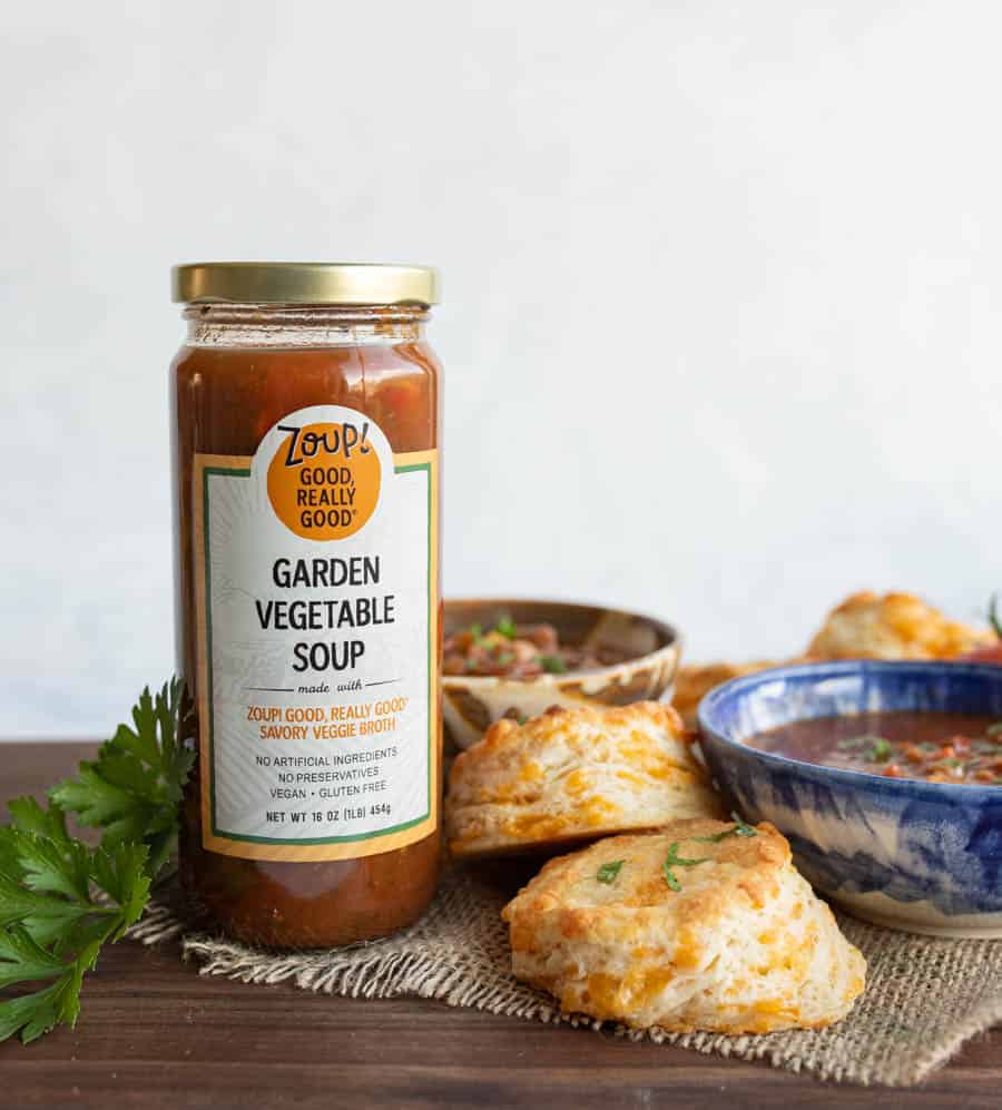 A jar of Zoup! Garden Vegetable Soup stands next to two blue bowls of soup and three golden, flaky cheddar biscuits on a woven placemat, with fresh parsley on the side.