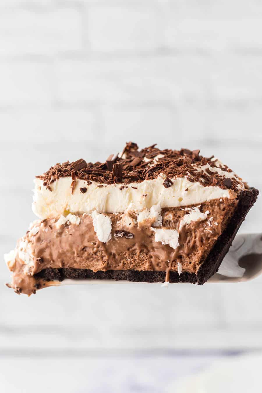 A slice of french silk pie with a chocolate cookie crust, creamy chocolate filling, whipped cream topping, and chocolate shavings on top, held up against a light, blurred background.