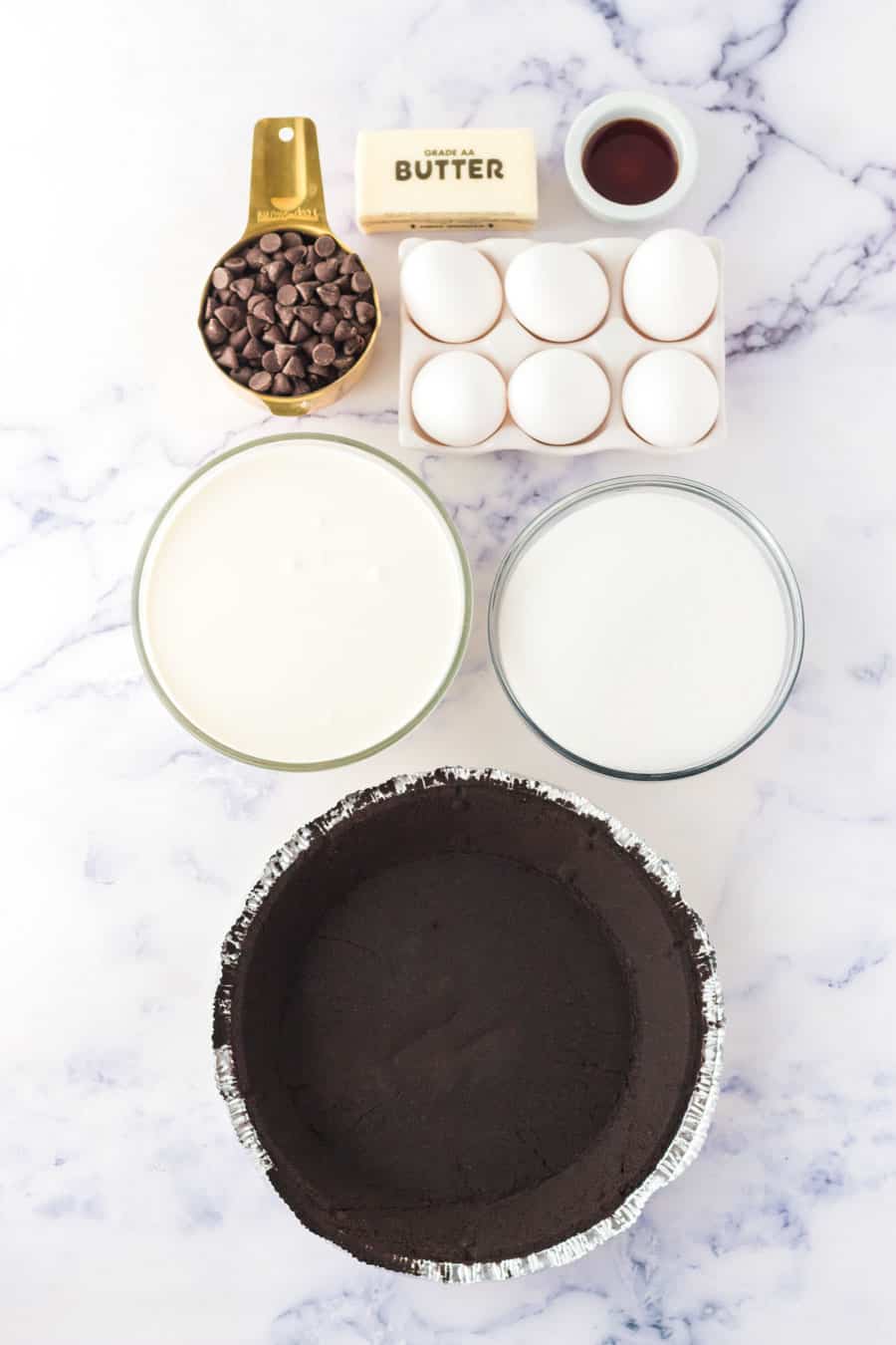 Ingredients for a classic french silk pie—chocolate pie crust, eggs, stick of butter, vanilla extract, chocolate chips, sugar, and cream—are arranged on a white marble surface.