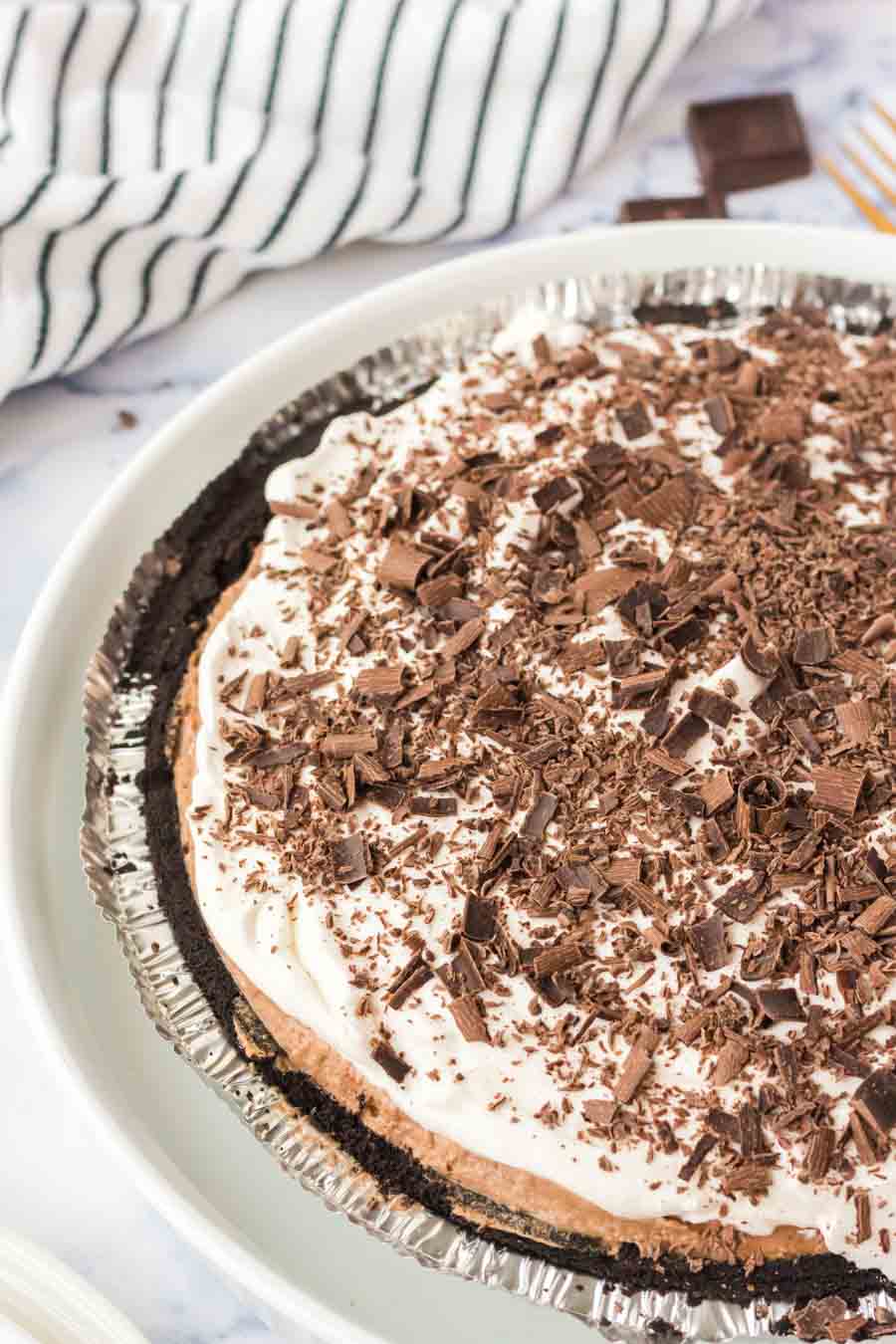A decadent french silk pie with a crumbly chocolate crust, topped with whipped cream and chocolate shavings, sits on a white plate. A striped cloth and chocolate pieces are visible in the background.