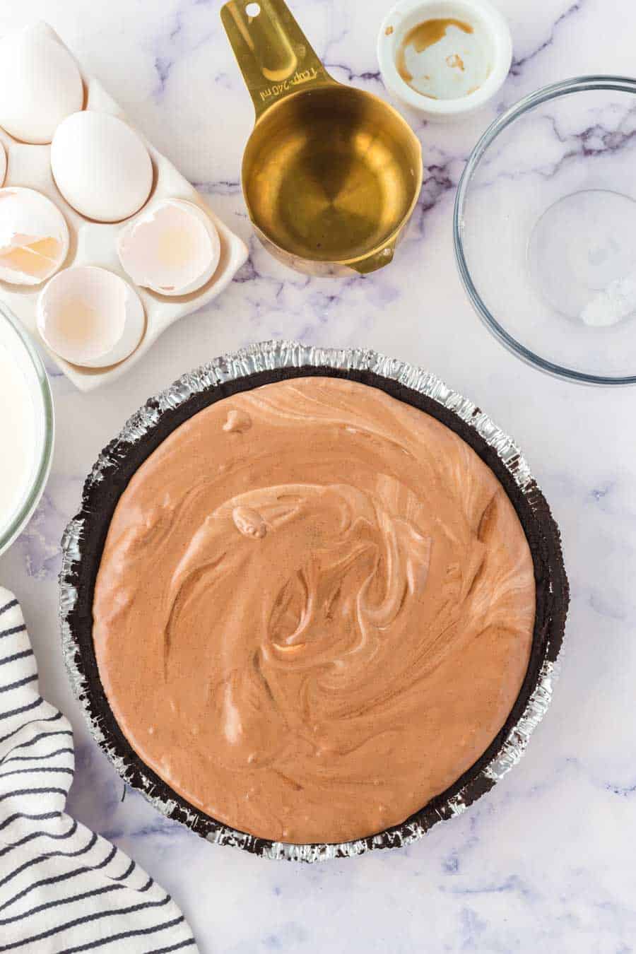 A french silk pie with a swirled top sits in a foil crust on a marble countertop, surrounded by a measuring cup, eggshells, a glass bowl, and a striped cloth.