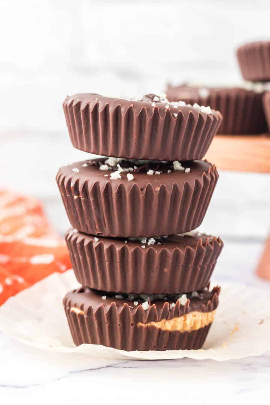 A stack of four homemade peanut butter cups with a sprinkle of sea salt on top, one with a bite taken out revealing the creamy peanut butter filling, sits on a white surface with a red cloth in the background.