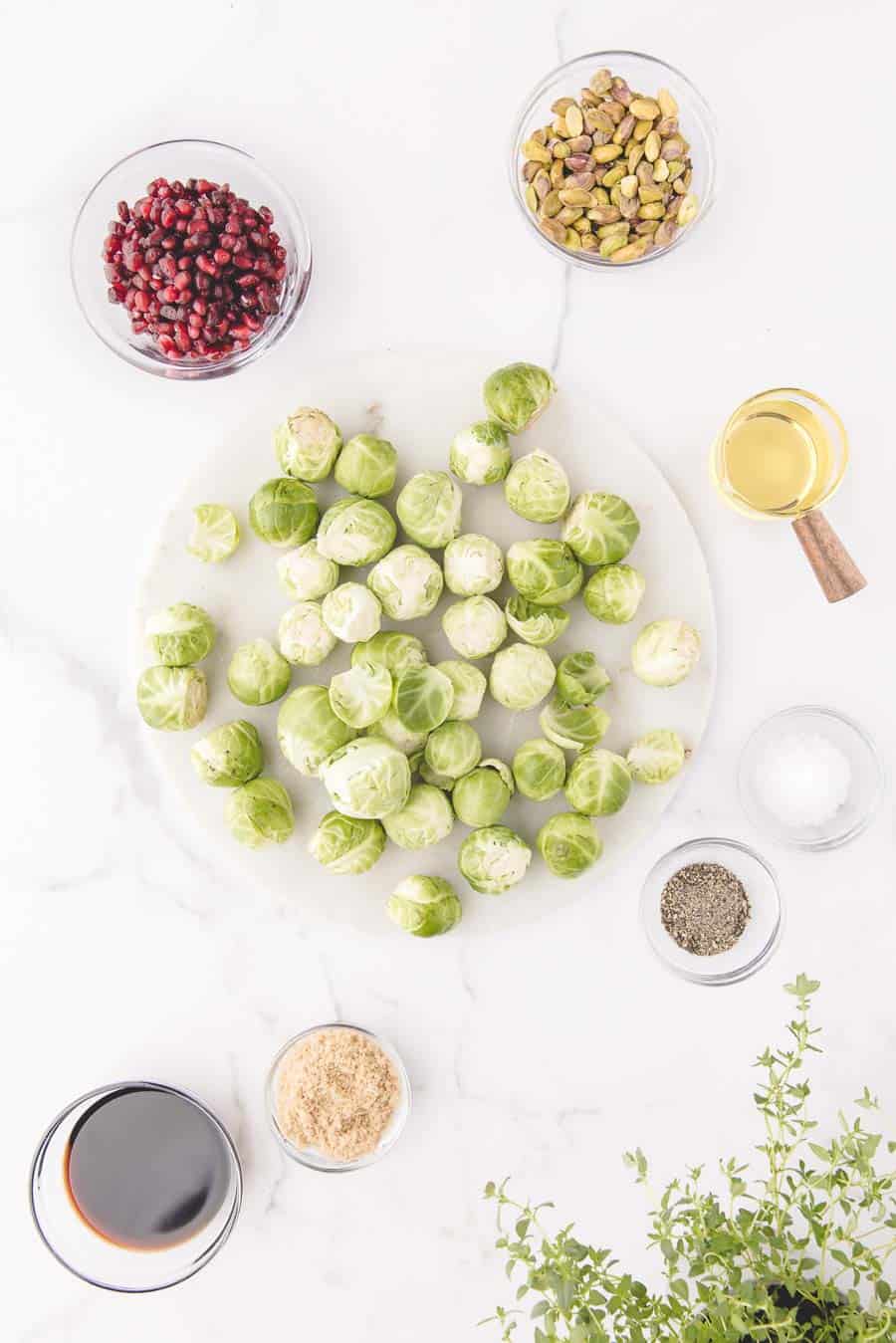 Overhead view of raw Brussels sprouts on a round white board, surrounded by small bowls of pomegranate seeds, pistachios, olive oil, and spices—perfect prep for making roasted Brussels sprouts in the oven with balsamic vinegar and fresh thyme.