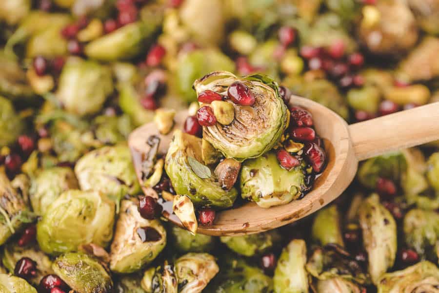 A wooden spoon holds roasted Brussels sprouts in the oven, topped with pomegranate seeds and pistachios, surrounded by more roasted Brussels sprouts in the background.