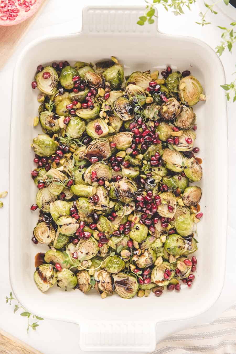 A white baking dish filled with roasted Brussels sprouts in the oven, garnished with pomegranate seeds and chopped pistachios. Fresh herbs are sprinkled on top, adding color and texture.