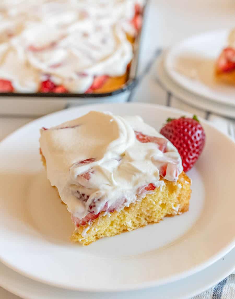 A slice of cake with whipped cream and strawberries on top, served on a white plate, next to a whole strawberry. A tray with more cake is blurred in the background.