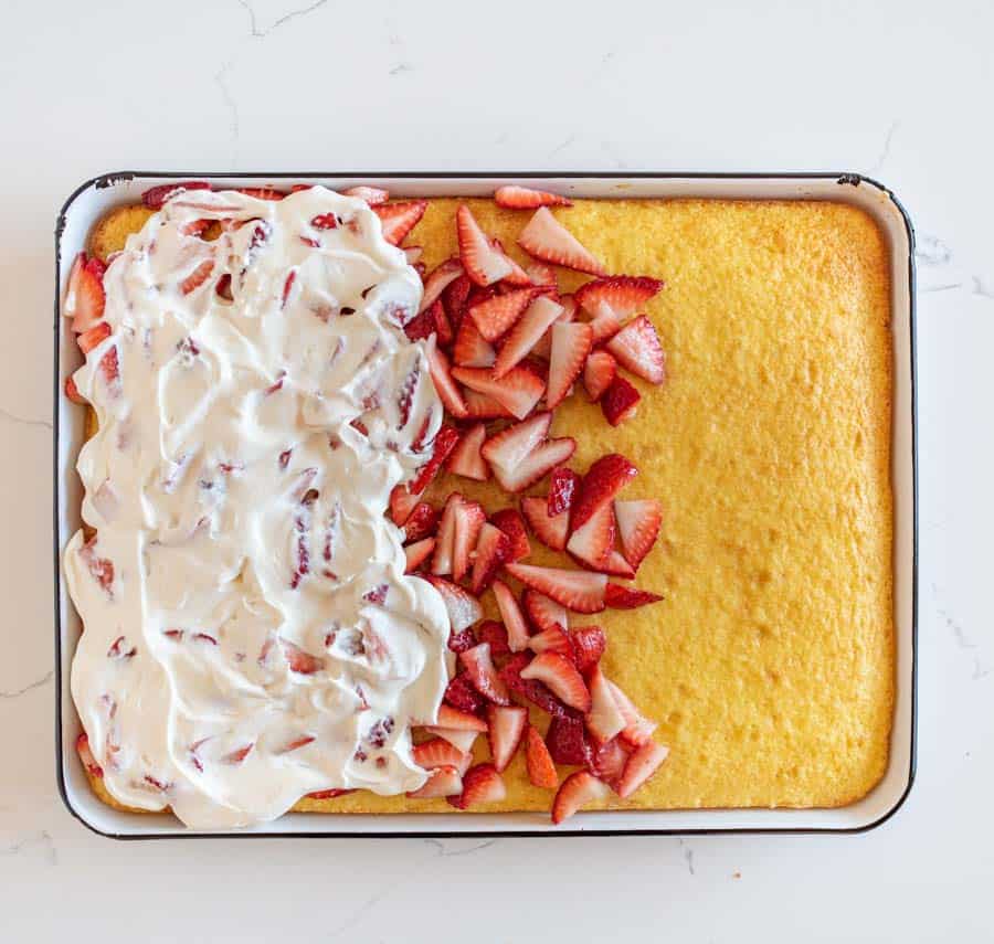A rectangular cake, resembling an easy strawberry shortcake cake recipe, is partially topped with whipped cream and sliced strawberries on the left side. The right side remains plain. Served on a white dish, it sits elegantly atop a marble countertop.