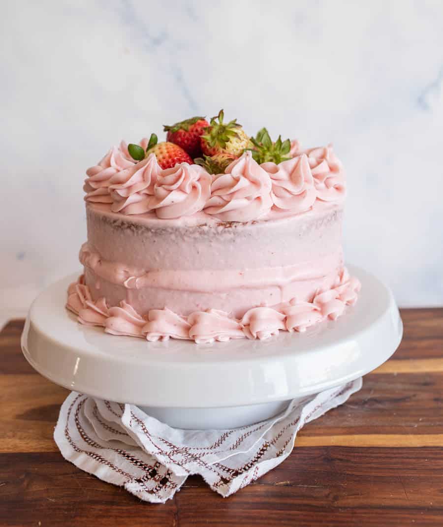 A pink frosted cake, inspired by a classic strawberry cake recipe, is decorated with swirls of icing and fresh strawberries on top, displayed on a white cake stand with a lace cloth, set on a wooden surface.