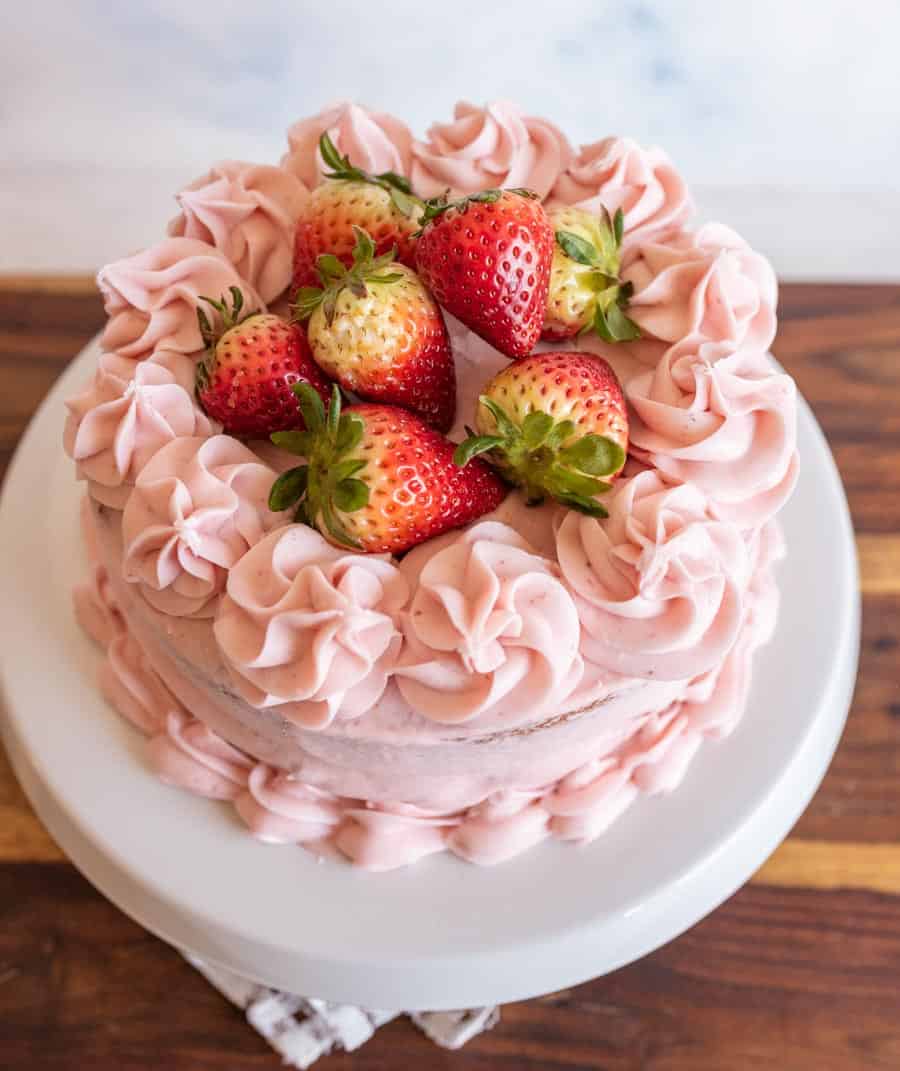 A round strawberry cake recipe with pink frosting and decorative swirls, topped with fresh whole strawberries, sits on a white cake stand on a wooden surface.