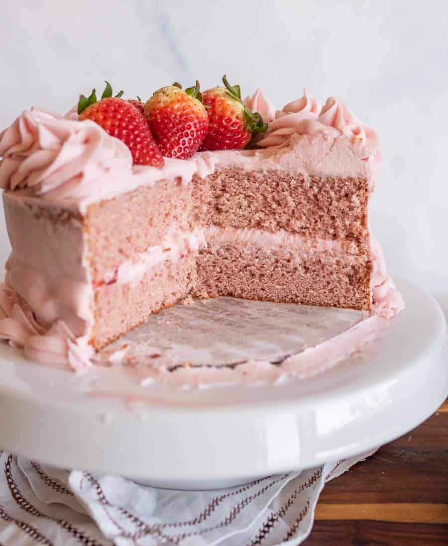 A pink frosted strawberry layer cake from a favorite strawberry cake recipe, with a slice missing, is topped with fresh strawberries and sits on a white cake stand over a cloth napkin.