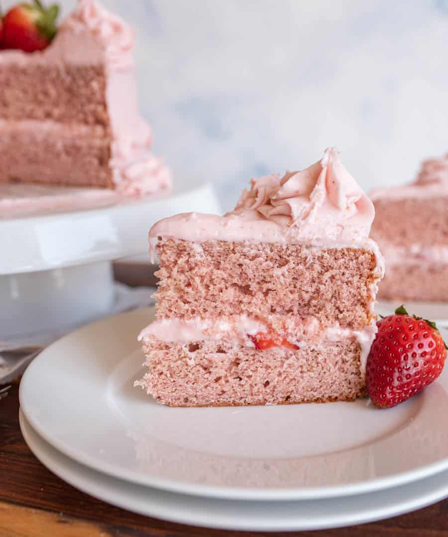 A slice of pink-layered strawberry cake with pink frosting and a fresh strawberry on the side, placed on a white plate. The rest of this tempting strawberry cake recipe sits in the background.