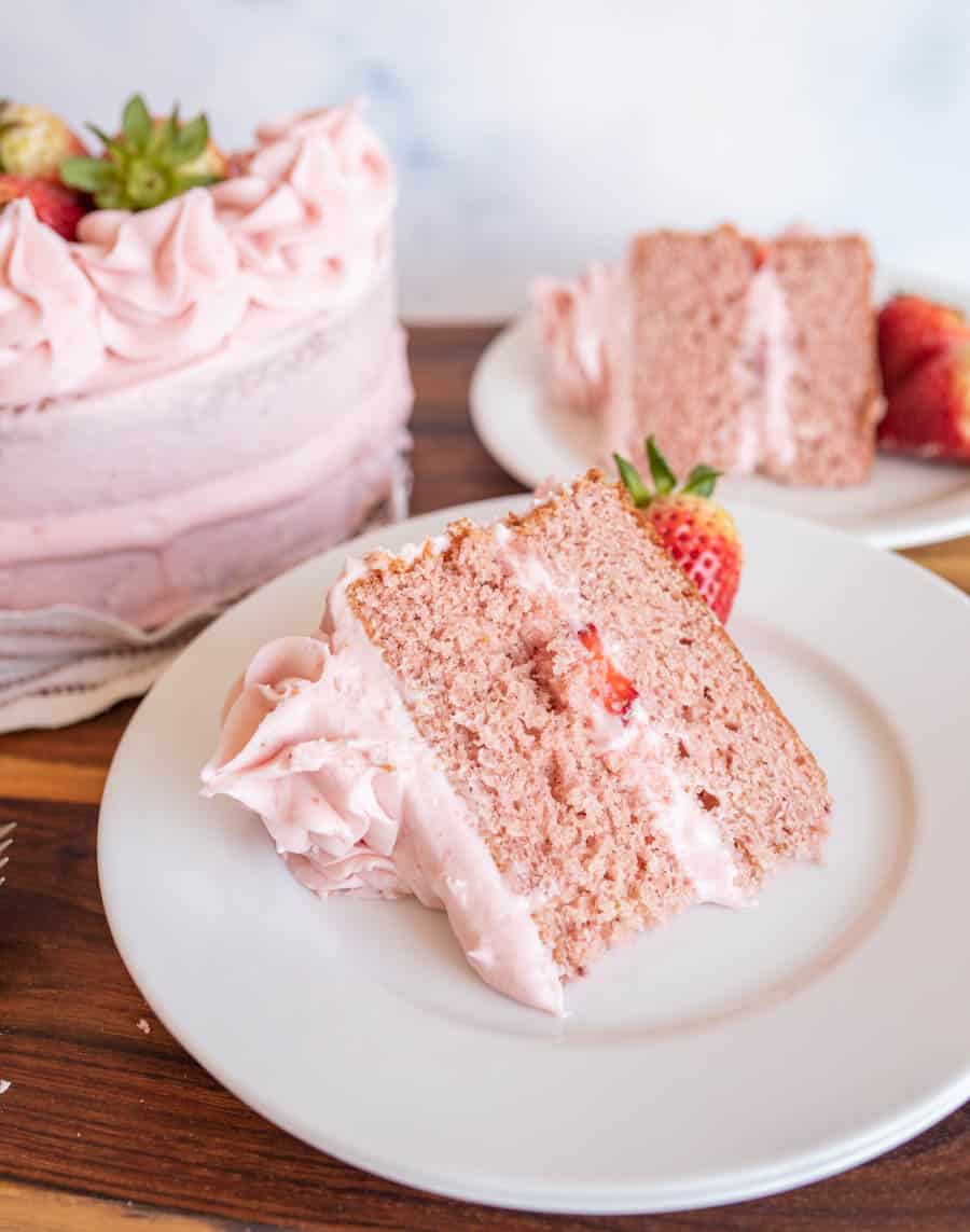 A slice of pink strawberry cake with strawberry frosting sits on a white plate, garnished with a fresh strawberry. The whole cake and another slice are visible in the background—perfect inspiration for your next strawberry cake recipe.
