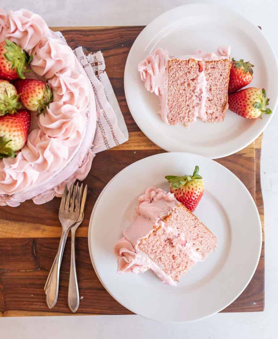 A pink frosted strawberry cake recipe on a wooden board, with two slices served on white plates, each garnished with a whole strawberry and extra frosting. Three forks are placed next to the plates.