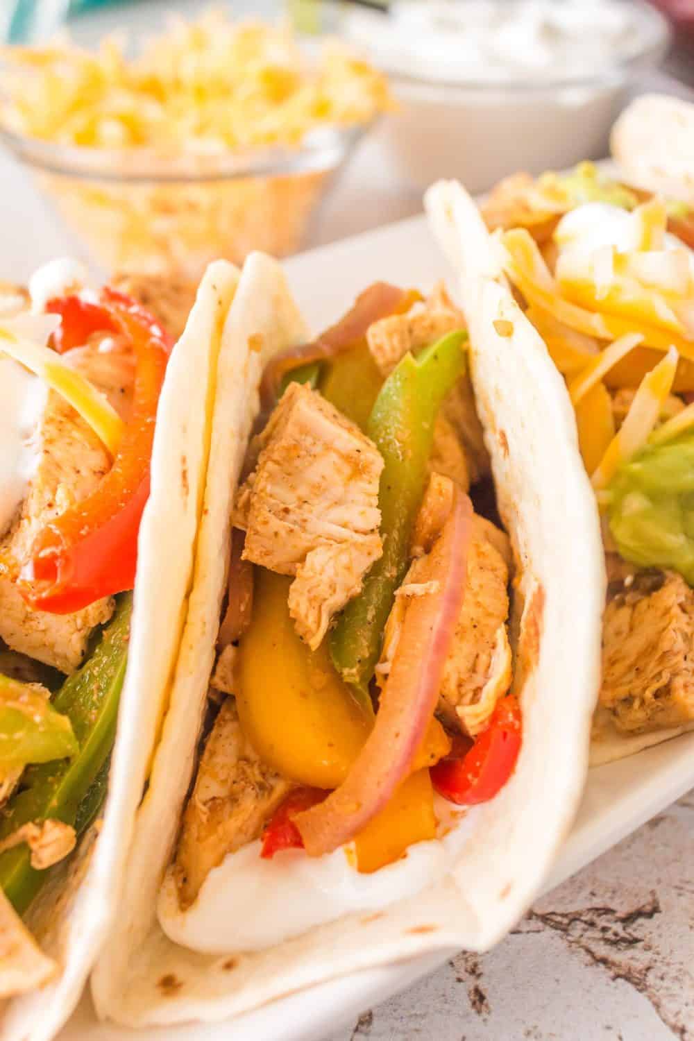 Three soft flour tortillas filled with seasoned sheet pan chicken fajitas, sliced bell peppers, red onions, melted cheese, guacamole, and sour cream, served on a white plate with toppings in the background.