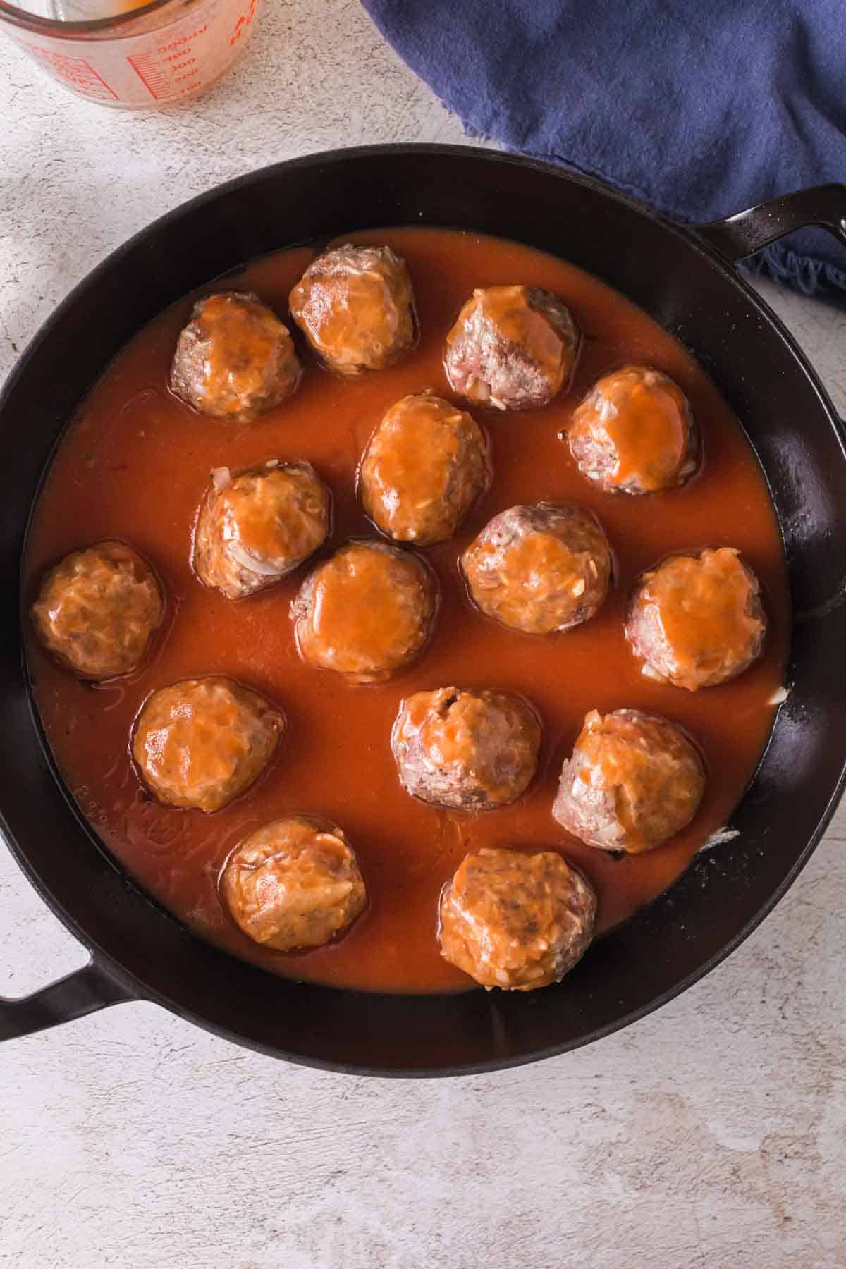A black skillet filled with porcupine meatballs covered in red tomato sauce, sitting on a light-colored surface with a blue cloth and a glass measuring cup partially visible nearby.