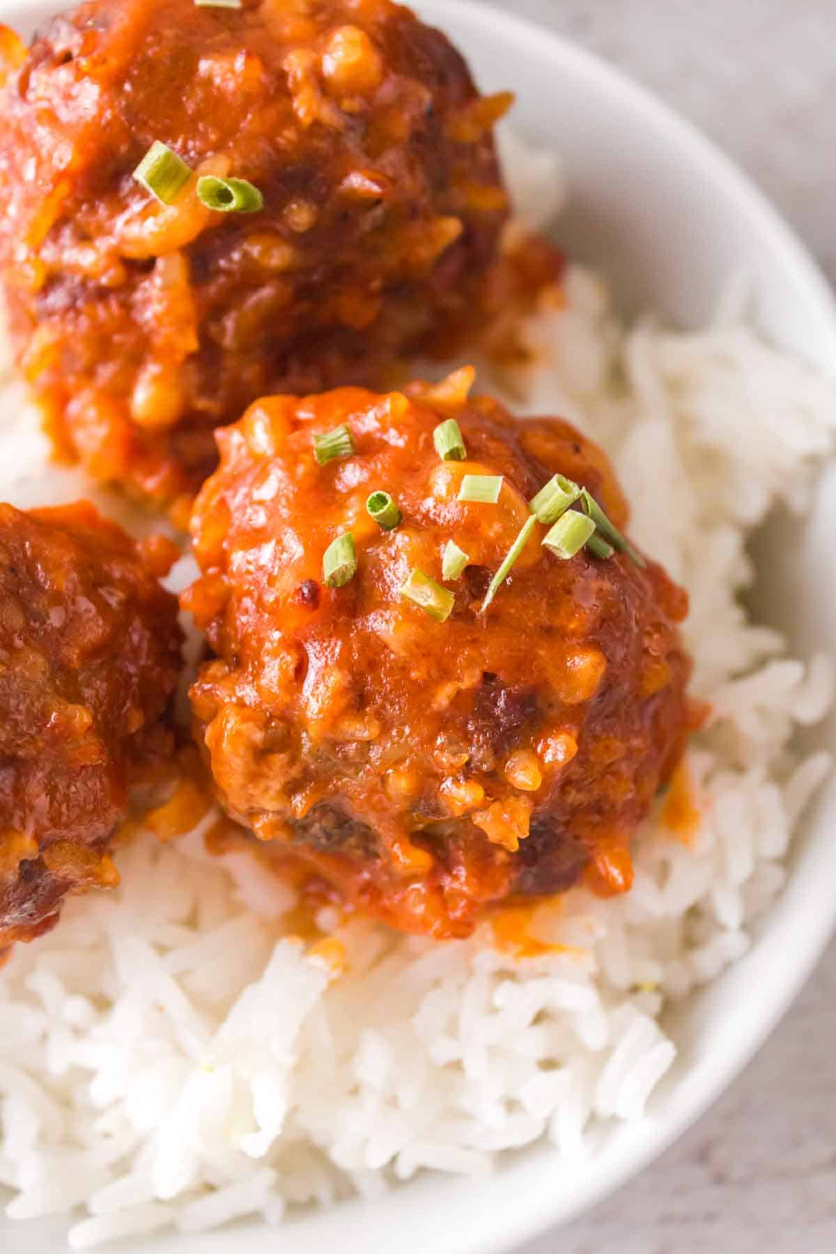 Three saucy porcupine meatballs topped with chopped green herbs are served on a bed of white rice in a white bowl.