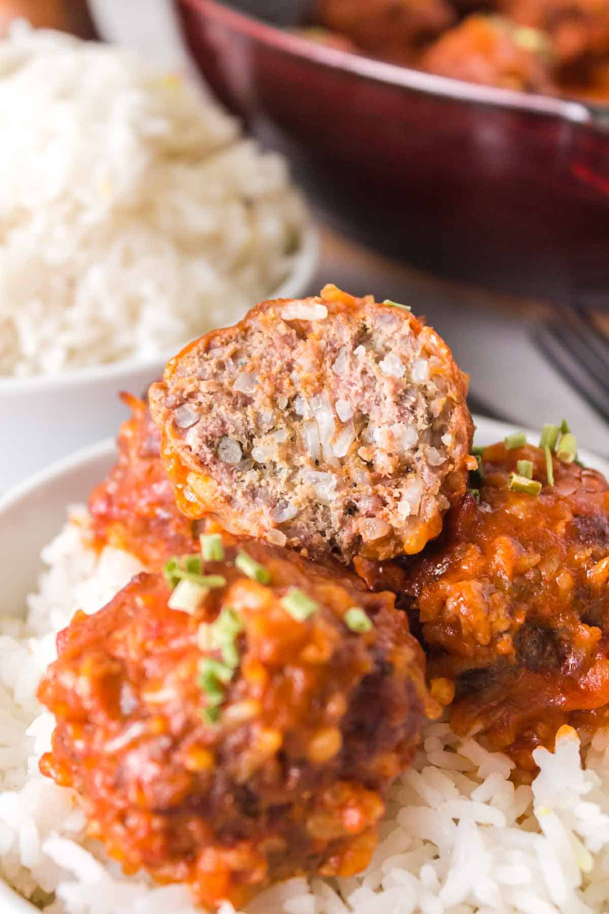 Three porcupine meatballs with tomato sauce served over white rice, with one meatball cut in half to show its texture. The dish is garnished with chopped green herbs, and more rice is visible in the background.