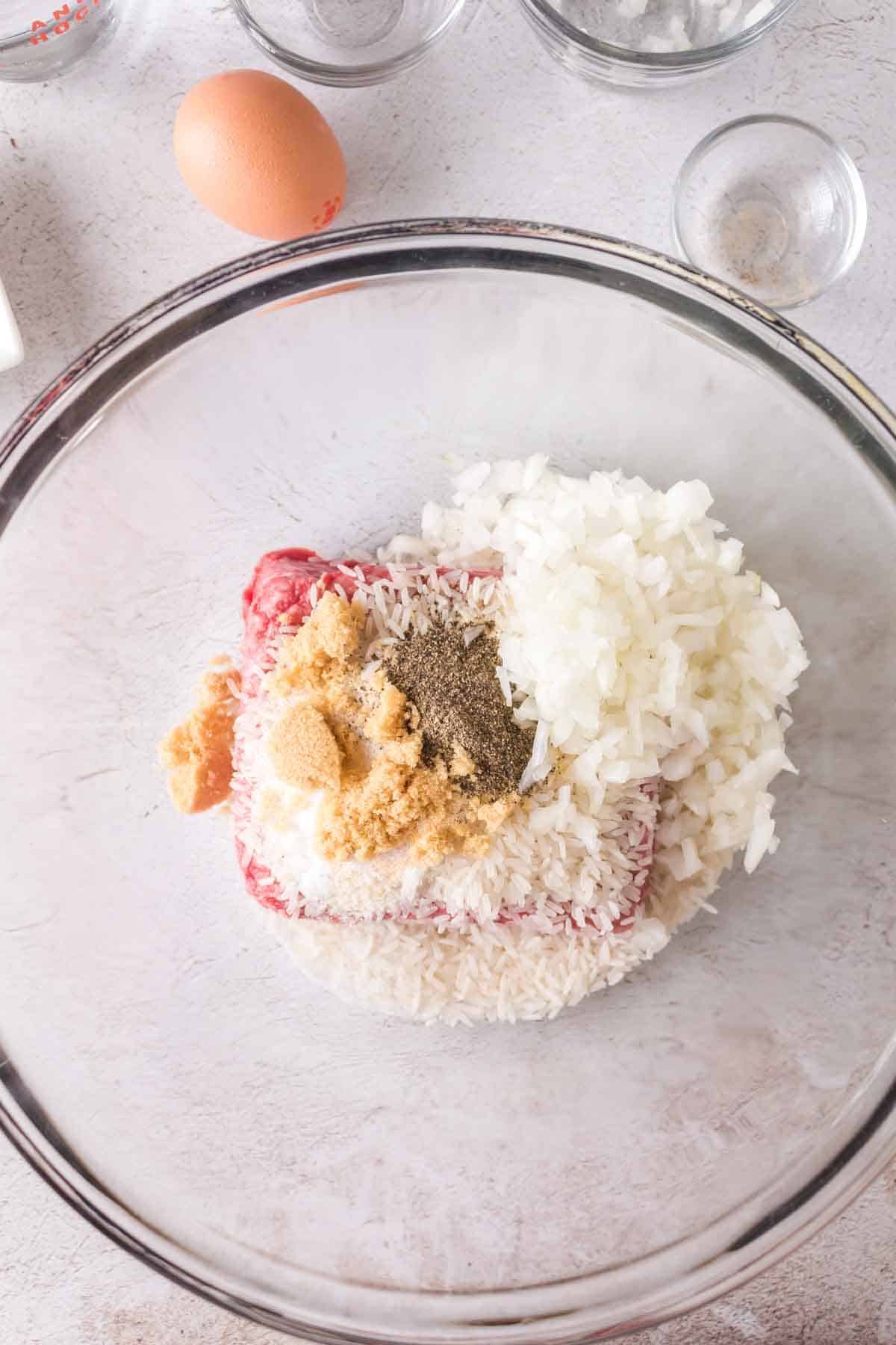 A glass bowl containing ground meat, chopped onions, uncooked rice, an egg, and seasonings—ingredients for porcupine meatballs—ready to be mixed. Surrounding the bowl are empty glass containers on a light surface.