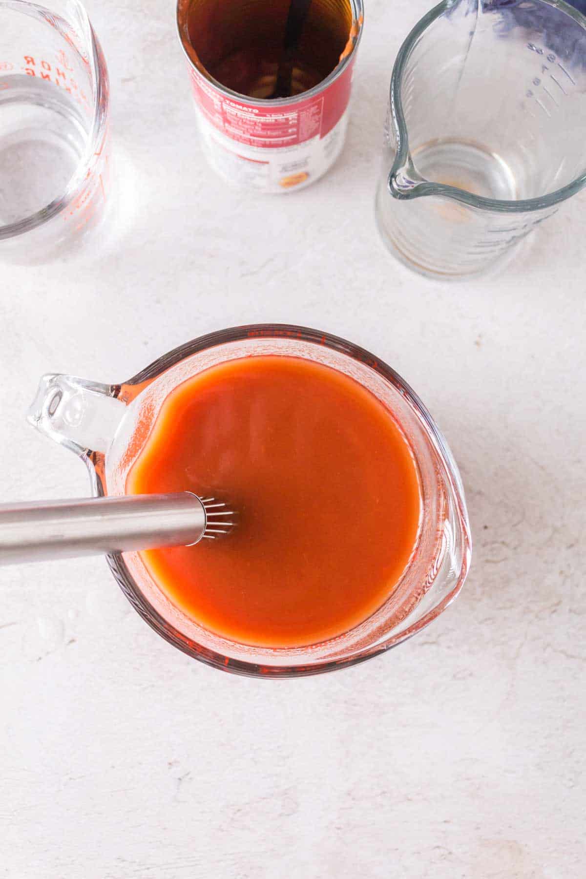 A metal whisk mixes a reddish-orange liquid in a glass measuring cup on a light surface—perfect for preparing sauce for porcupine meatballs—with an empty measuring cup, an open can, and a cup of water nearby.