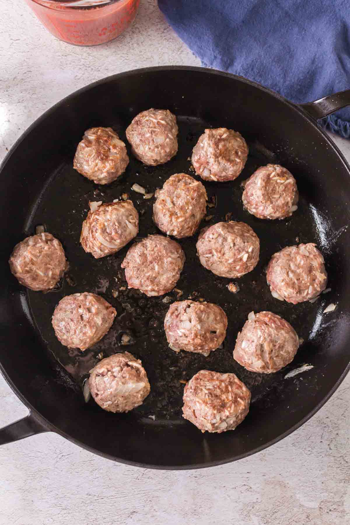 A black skillet with fourteen uncooked or lightly browned porcupine meatballs arranged inside, set on a light-colored surface next to a blue cloth and a partial view of a jar with red sauce.