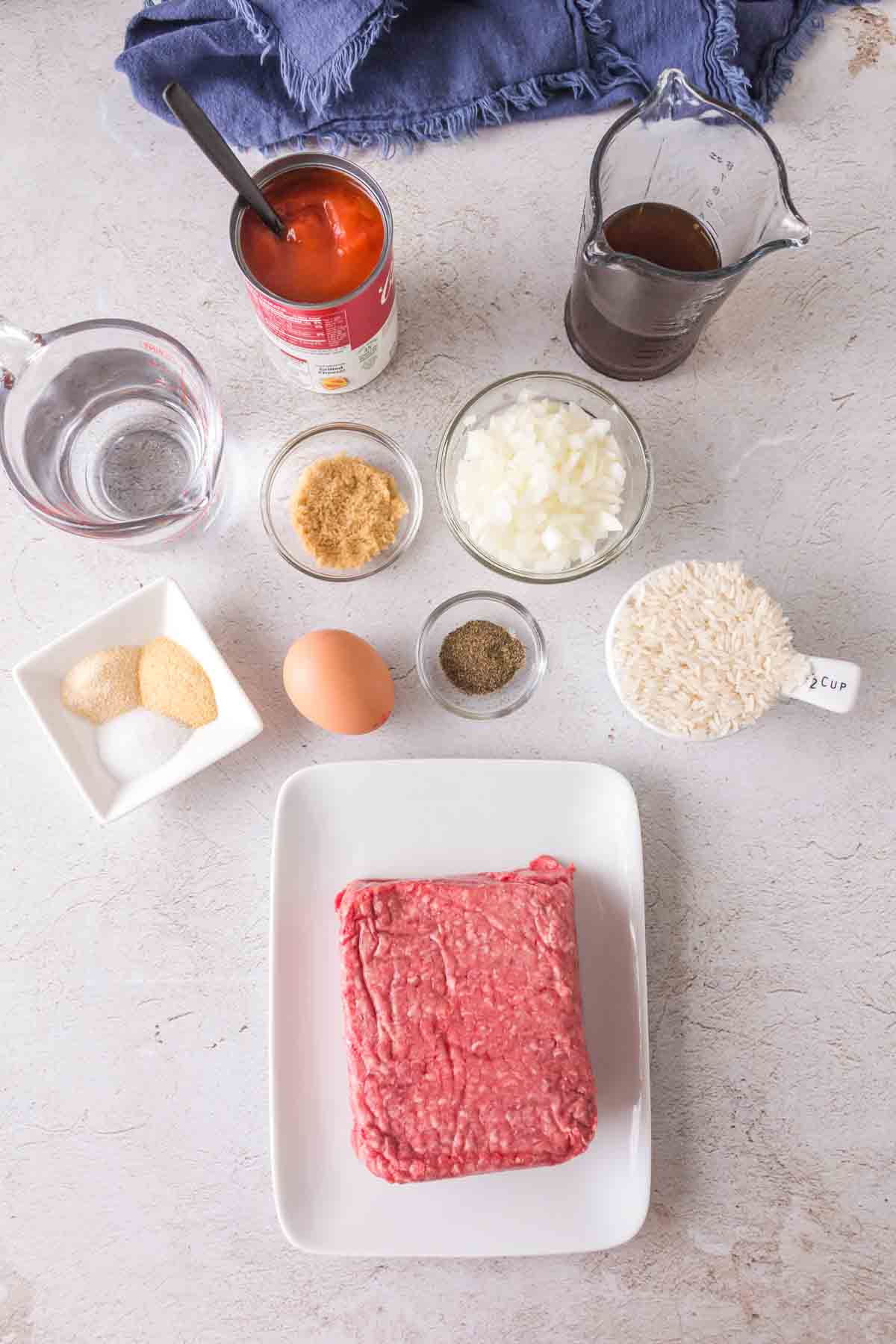 Raw ground beef on a white plate, surrounded by small bowls of chopped onion, rice, seasonings, and other essentials for making classic porcupine meatballs on a light countertop with a blue cloth napkin.