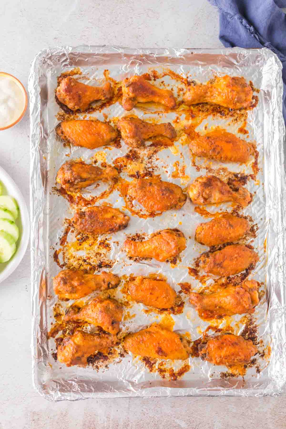 A baking tray lined with foil holds rows of golden-brown hot wings with crispy skin. A small bowl of dipping sauce and cucumber slices are visible to the side on a white plate. A dark blue cloth is partially visible in the corner.